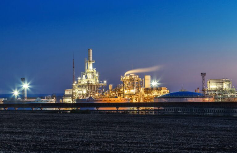 Oil refinery in Burgas, Bulgaria at night. Image shows white structure consisting of chimneys spouting emissions glowing with night lighting and a dark blue night sky behind it.
